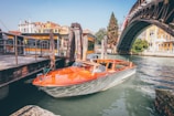 A classic wooden speedboat is moored at a dock along a canal. The boat has an elegant design with a polished wooden deck and a sleek white hull. Adjacent to the boat, there's a wooden pier with weathered posts and chains. In the background, a large arched iron bridge spans the canal, with colorful historic buildings lining the waterfront.