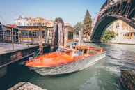 A classic wooden speedboat is moored at a dock along a canal. The boat has an elegant design with a polished wooden deck and a sleek white hull. Adjacent to the boat, there's a wooden pier with weathered posts and chains. In the background, a large arched iron bridge spans the canal, with colorful historic buildings lining the waterfront.