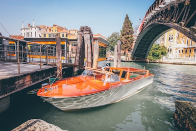 A classic wooden speedboat is moored at a dock along a canal. The boat has an elegant design with a polished wooden deck and a sleek white hull. Adjacent to the boat, there's a wooden pier with weathered posts and chains. In the background, a large arched iron bridge spans the canal, with colorful historic buildings lining the waterfront.
