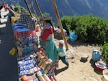 A market stall is set up along a paved path with local people dressed in traditional clothing. The stall displays a colorful assortment of snacks, drinks, and fresh bananas. Behind the market setup, lush green bushes and a mountainous landscape are visible. A young child stands beside a woman, smiling.