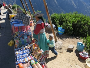 A market stall is set up along a paved path with local people dressed in traditional clothing. The stall displays a colorful assortment of snacks, drinks, and fresh bananas. Behind the market setup, lush green bushes and a mountainous landscape are visible. A young child stands beside a woman, smiling.