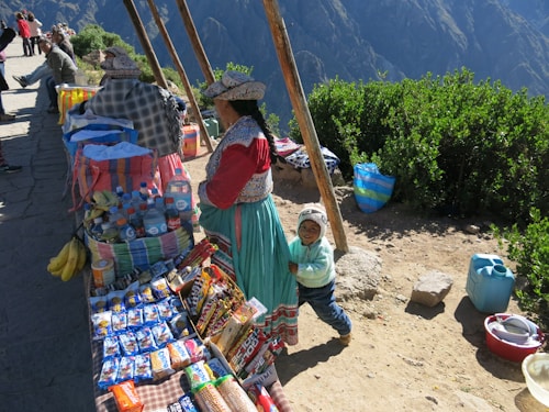 A market stall is set up along a paved path with local people dressed in traditional clothing. The stall displays a colorful assortment of snacks, drinks, and fresh bananas. Behind the market setup, lush green bushes and a mountainous landscape are visible. A young child stands beside a woman, smiling.