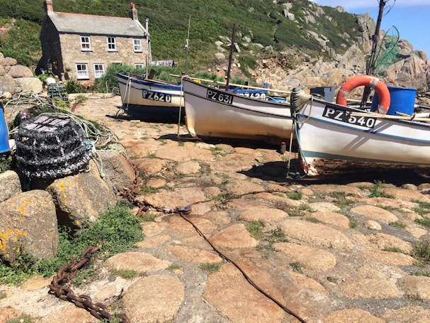 A quaint coastal scene featuring a small stone cottage with a tiled roof and white-framed windows. In front of the house, two boats with identification numbers rest on a rocky shoreline, accompanied by fishing nets and lobster pots. The landscape is adorned with lush greenery and rugged rocky outcrops under a clear blue sky.