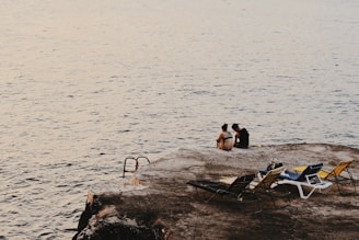 man and woman sitting on cliff beside sea and sun loungers