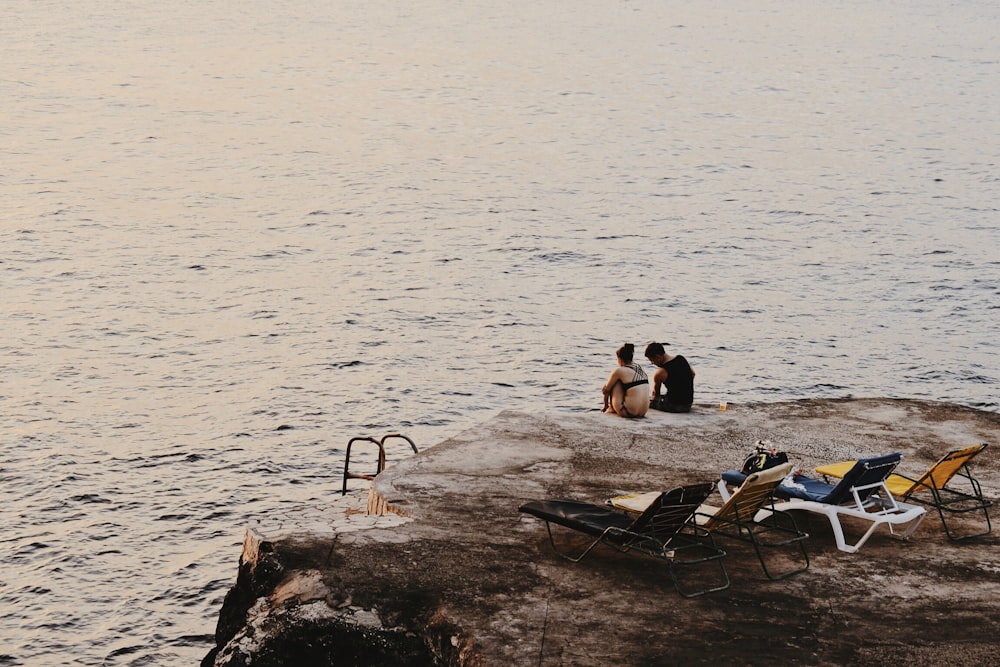 man and woman sitting on cliff beside sea and sun loungers