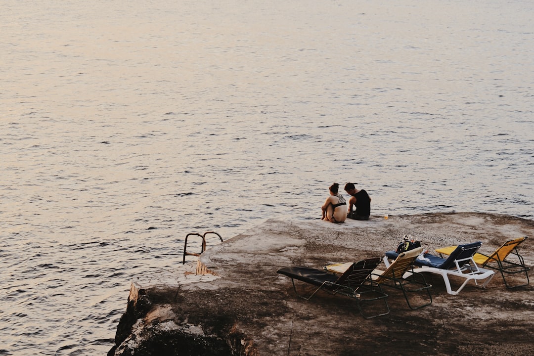 man and woman sitting on cliff beside sea and sun loungers,