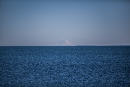 A vast expanse of calm ocean stretches toward the horizon, meeting a clear blue sky. In the distance, a snow-capped mountain rises above the horizon, adding a sense of majesty to the serene seascape.