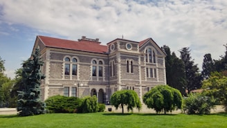A historic stone building with a red-tiled roof stands prominently surrounded by green trees and shrubs. The architecture features arched windows and detailed stonework, creating a sense of classical elegance. The front lawn is lush and well-maintained, and the sky above is partly cloudy, adding a serene backdrop.