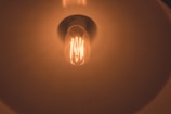 A glowing incandescent light bulb is visible from a close-up perspective. The design of the bulb's filament is distinct, and it is housed within a lamp with a reflective shade, which helps diffuse the warm, orange light emitted by the bulb.