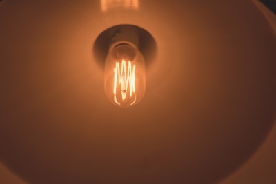 A glowing incandescent light bulb is visible from a close-up perspective. The design of the bulb's filament is distinct, and it is housed within a lamp with a reflective shade, which helps diffuse the warm, orange light emitted by the bulb.