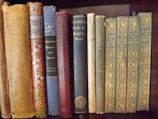A collection of vintage and classic books of various sizes and colors are neatly lined up on a wooden shelf. The spines show titles such as 'Aristotle's Rhetoric and Poetics', 'Twelfth Night', and 'Poems by E.B. Browning'. The books vary in condition, indicating they are well-read and possibly valuable.