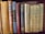 A collection of vintage and classic books of various sizes and colors are neatly lined up on a wooden shelf. The spines show titles such as 'Aristotle's Rhetoric and Poetics', 'Twelfth Night', and 'Poems by E.B. Browning'. The books vary in condition, indicating they are well-read and possibly valuable.