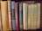 A collection of vintage and classic books of various sizes and colors are neatly lined up on a wooden shelf. The spines show titles such as 'Aristotle's Rhetoric and Poetics', 'Twelfth Night', and 'Poems by E.B. Browning'. The books vary in condition, indicating they are well-read and possibly valuable.