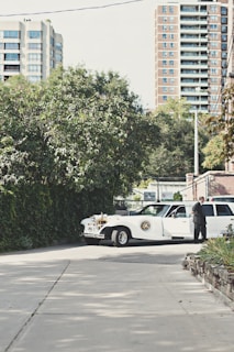 A vintage-style white limousine is parked on a quiet street, surrounded by greenery and with tall residential buildings in the background. A person in a suit stands next to the car, appearing to be engaged in some activity at the open car door.