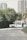 A vintage-style white limousine is parked on a quiet street, surrounded by greenery and with tall residential buildings in the background. A person in a suit stands next to the car, appearing to be engaged in some activity at the open car door.