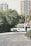 A vintage-style white limousine is parked on a quiet street, surrounded by greenery and with tall residential buildings in the background. A person in a suit stands next to the car, appearing to be engaged in some activity at the open car door.
