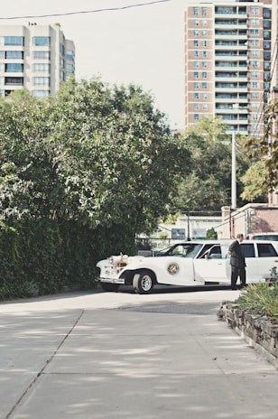 A vintage-style white limousine is parked on a quiet street, surrounded by greenery and with tall residential buildings in the background. A person in a suit stands next to the car, appearing to be engaged in some activity at the open car door.