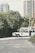 A vintage-style white limousine is parked on a quiet street, surrounded by greenery and with tall residential buildings in the background. A person in a suit stands next to the car, appearing to be engaged in some activity at the open car door.