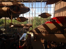 Several vintage and modern airplanes are displayed indoors in a museum with large windows allowing natural light to illuminate the space. The airplanes are suspended from the ceiling, showcasing different eras of aviation history.