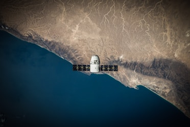 A satellite is positioned over a coastal landscape, highlighting a stark contrast between the deep blue ocean and the arid, brown rocky terrain. The satellite features a large solar panel array and is centered in the image, capturing both natural and technological elements.