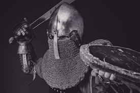 A medieval knight wearing a metal helmet and chainmail, holding a sword raised in one hand and a weathered shield in the other, poised in an action stance against a dark background.