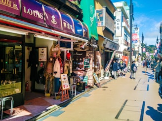 A busy street lined with shops and boutiques showing various clothing items on display outside. People are walking along the sidewalk, and some are stopping to look at the stores. The storefronts feature colorful signage, including awnings and banners. A bright, clear sky and shadows from the buildings indicate sunlight and daytime.