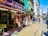 A busy street lined with shops and boutiques showing various clothing items on display outside. People are walking along the sidewalk, and some are stopping to look at the stores. The storefronts feature colorful signage, including awnings and banners. A bright, clear sky and shadows from the buildings indicate sunlight and daytime.