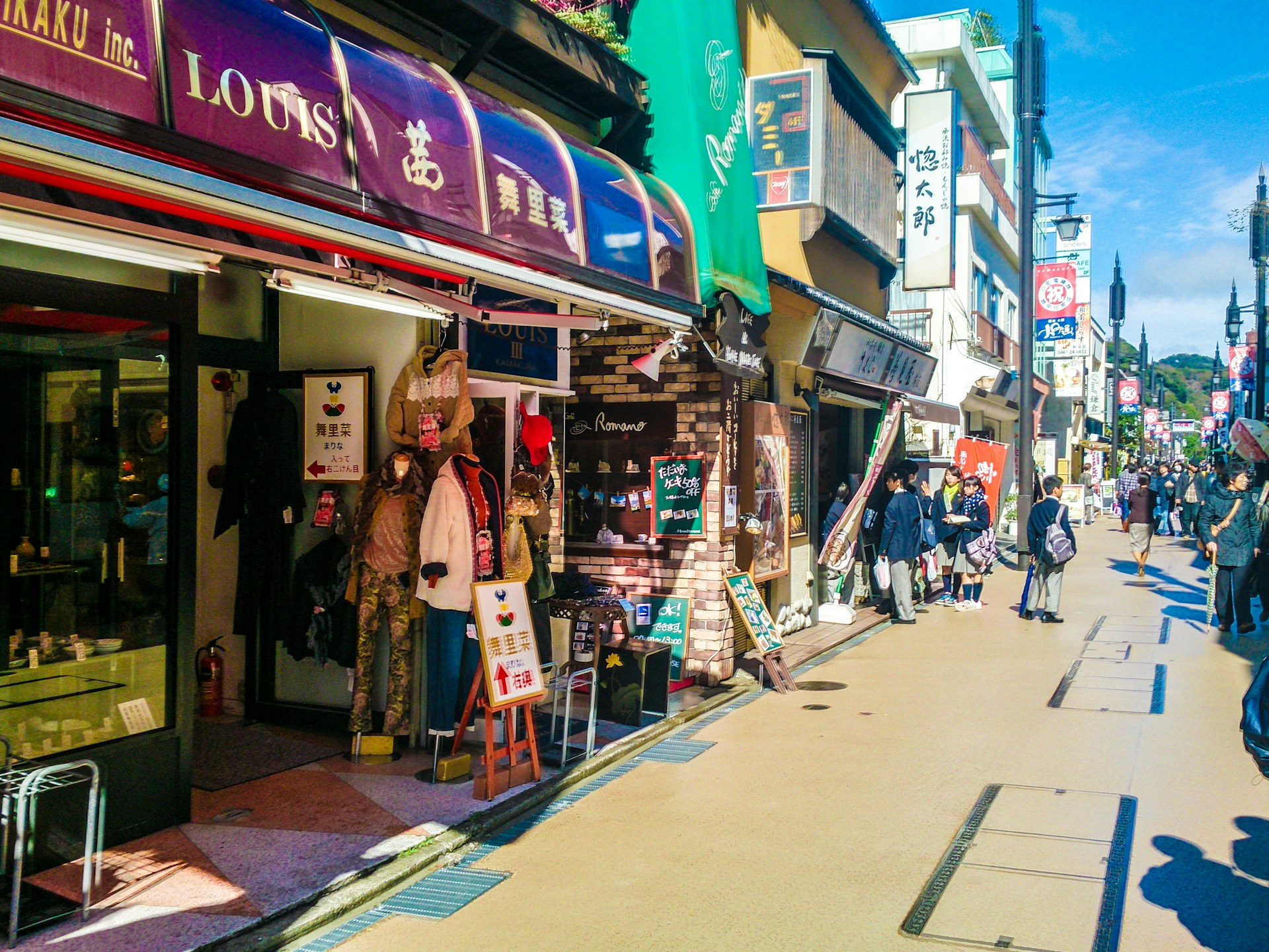 A busy street lined with shops and boutiques showing various clothing items on display outside. People are walking along the sidewalk, and some are stopping to look at the stores. The storefronts feature colorful signage, including awnings and banners. A bright, clear sky and shadows from the buildings indicate sunlight and daytime.