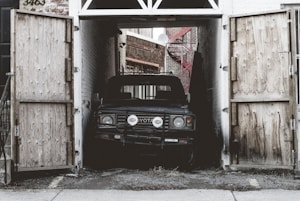 A vintage Toyota vehicle is parked inside a narrow garage with wooden doors that are open. The scene portrays an urban environment with brick walls and a red external staircase leading up one of the buildings.