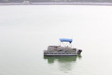 A pontoon boat with a blue canopy floats on a calm, light-green lake. The water is still, reflecting the overcast sky. The distant shoreline and trees are faintly visible in the background.