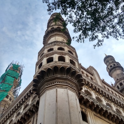 A detailed view of a historical monument showcasing intricate architectural details. Tall towers with ornate designs are visible, framed by a blue sky. Some parts of the structure are covered with green scaffolding, indicating restoration work. The perspective gives a sense of grandeur and height.
