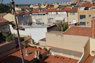 Residential area with multiple buildings featuring terracotta roofs and light-colored walls. The setting includes an urban landscape with a mix of small balconies and urban gardens adorned with potted plants. Rooftop terraces can be seen, along with a clear blue sky in the background. The area appears densely built with closely packed houses.