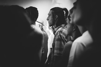 A black and white photograph captures a group of people standing closely together, likely in a crowded setting. The central figure is a person with long hair tied back, wearing a plaid shirt. The faces of the individuals are lit from the side, creating a dramatic and contemplative atmosphere.