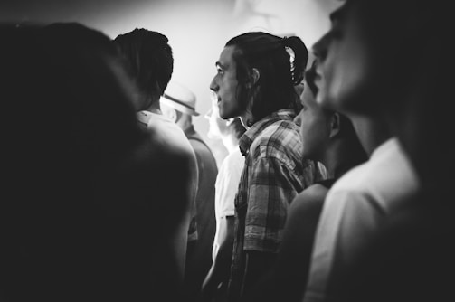 A black and white photograph captures a group of people standing closely together, likely in a crowded setting. The central figure is a person with long hair tied back, wearing a plaid shirt. The faces of the individuals are lit from the side, creating a dramatic and contemplative atmosphere.