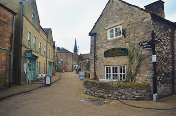 A quaint village street lined with old stone buildings. On the right, a charming cottage-style bakery with a stone facade and ivy growing up the walls. On the left, a cozy cafe with outdoor seating visible on the sidewalk. The street appears quiet and is made of cobblestones, leading towards a distant church spire in the background.
