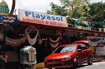 A souvenir and gift shop with a sign reading 'Playasol Souvenirs & Gift Shop', offering various items such as hanging hammocks and T-shirts. A bright red car is parked in front of the store. Colorful flags and greenery surround the shop.