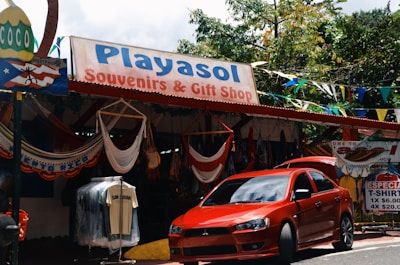 A souvenir and gift shop with a sign reading 'Playasol Souvenirs & Gift Shop', offering various items such as hanging hammocks and T-shirts. A bright red car is parked in front of the store. Colorful flags and greenery surround the shop.