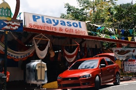 A souvenir and gift shop with a sign reading 'Playasol Souvenirs & Gift Shop', offering various items such as hanging hammocks and T-shirts. A bright red car is parked in front of the store. Colorful flags and greenery surround the shop.