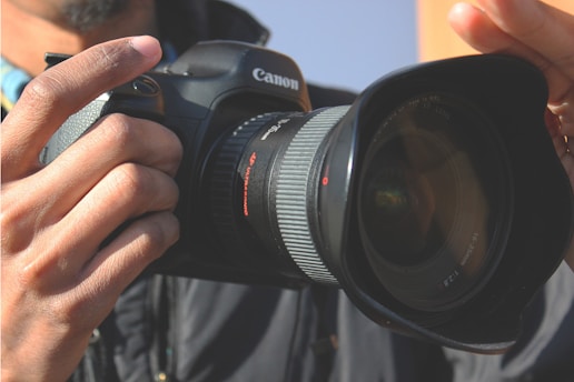 A person holding a Canon DSLR camera with a large lens. The camera is being held with both hands, focusing on the details of the lens and the camera body. The scene suggests the person is about to take a photograph.