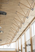 The image depicts the interior of a modern building featuring a high wooden ceiling supported by a series of angled wooden beams connected with metal joints and rods. Large windows on the right allow abundant natural light to illuminate the space. Various light fixtures and HVAC units are mounted on the ceiling and walls.