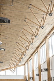 The image depicts the interior of a modern building featuring a high wooden ceiling supported by a series of angled wooden beams connected with metal joints and rods. Large windows on the right allow abundant natural light to illuminate the space. Various light fixtures and HVAC units are mounted on the ceiling and walls.
