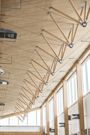 The image depicts the interior of a modern building featuring a high wooden ceiling supported by a series of angled wooden beams connected with metal joints and rods. Large windows on the right allow abundant natural light to illuminate the space. Various light fixtures and HVAC units are mounted on the ceiling and walls.