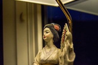 A porcelain figurine of a traditional female figure holds a decorated fan and is adorned with detailed clothing and a flower in her hair. The lighting casts a warm glow on the figure, set against a dimly lit background.