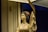 A porcelain figurine of a traditional female figure holds a decorated fan and is adorned with detailed clothing and a flower in her hair. The lighting casts a warm glow on the figure, set against a dimly lit background.