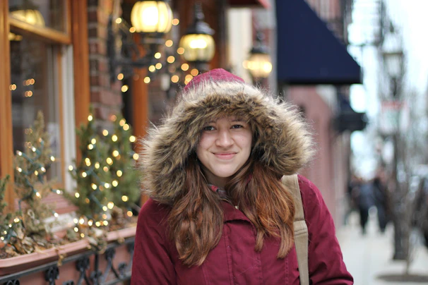 A young person wrapped in a cozy red scarf, standing against a snowy urban backdrop at dusk.