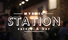 The image features the entrance of a restaurant or bar with a glass door displaying the words 'Mystic Station eatery & bar'. The background is slightly blurred, indicating an indoor setting with people sitting and ambient lighting from several bulbs creating a warm atmosphere.
