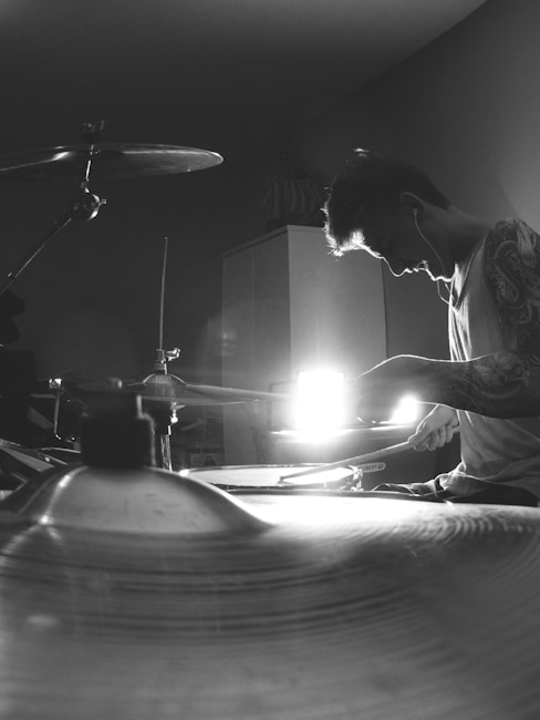 A person with earbuds is focused intently while playing a drum set. The image is taken in black and white, casting a dramatic contrast between light and shadow. The light source behind the person creates a striking silhouette effect. Visible are parts of a drum kit, including cymbals and drumsticks in motion.