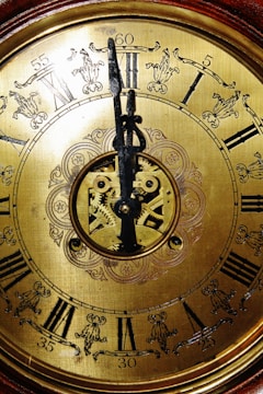 A close-up view of an ornate vintage clock face featuring Roman numerals. The intricate details include floral engravings and visible gears at the center, demonstrating craftsmanship. The metal hands are prominently displayed over a golden background, adding to the antique aesthetic.