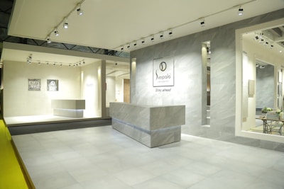An interior space featuring a modern showroom with large ceramic tiles and bright lighting. The walls display brand signage and various tile samples. A sleek, minimalist desk is centered, surrounded by a clean and spacious environment.