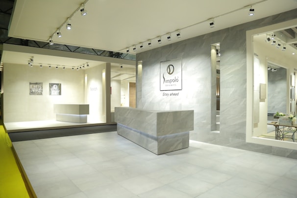 An interior space featuring a modern showroom with large ceramic tiles and bright lighting. The walls display brand signage and various tile samples. A sleek, minimalist desk is centered, surrounded by a clean and spacious environment.